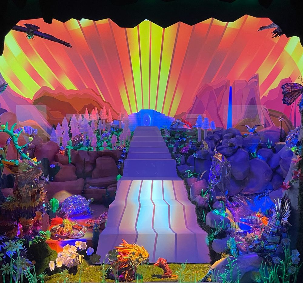 A diorama case is lit up in dayglo bright colors on a digital display. Many California animals cram into the foreground; some appear made of paper mache like piñatas while others light up inside. A staircase leads from the front of the diorama to the back directly down the middle. At the top of the stairs is a door. The sky above the door is lit up like sunrise.