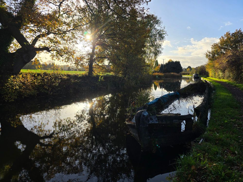 Sunlight streams through autumn trees over a dark, calm canal with an old boat moored to the bank