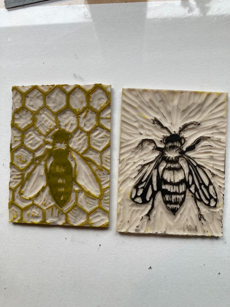 Two carved lino printing blocks. The one on the left is inked in yellow and the one on the right is inked in black. The layers combine to create a print of black bee on yellow honeycomb
