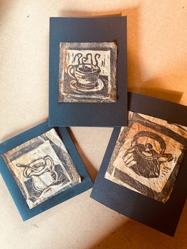 Three small linoprint images printed onto used teabags. Each print is pasted onto a black card. The images are 'tea' themed, with a steaming cup of tea, a jug, and a kettle.