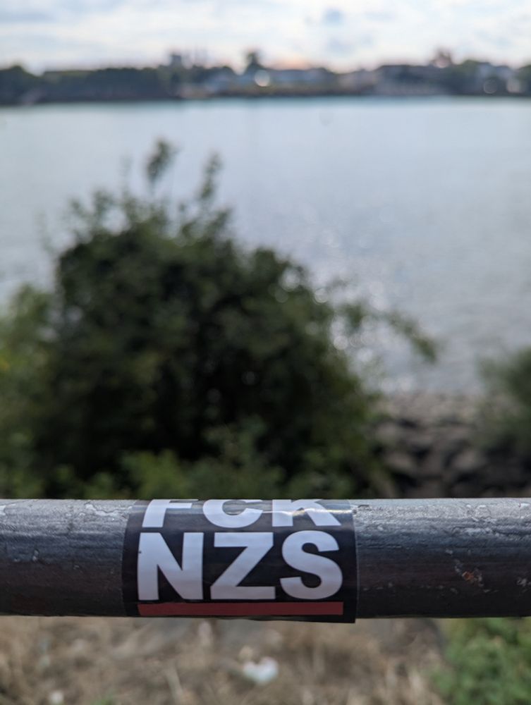 Fck nzs Sticker auf einem Geländer, dahinter der Rhein 