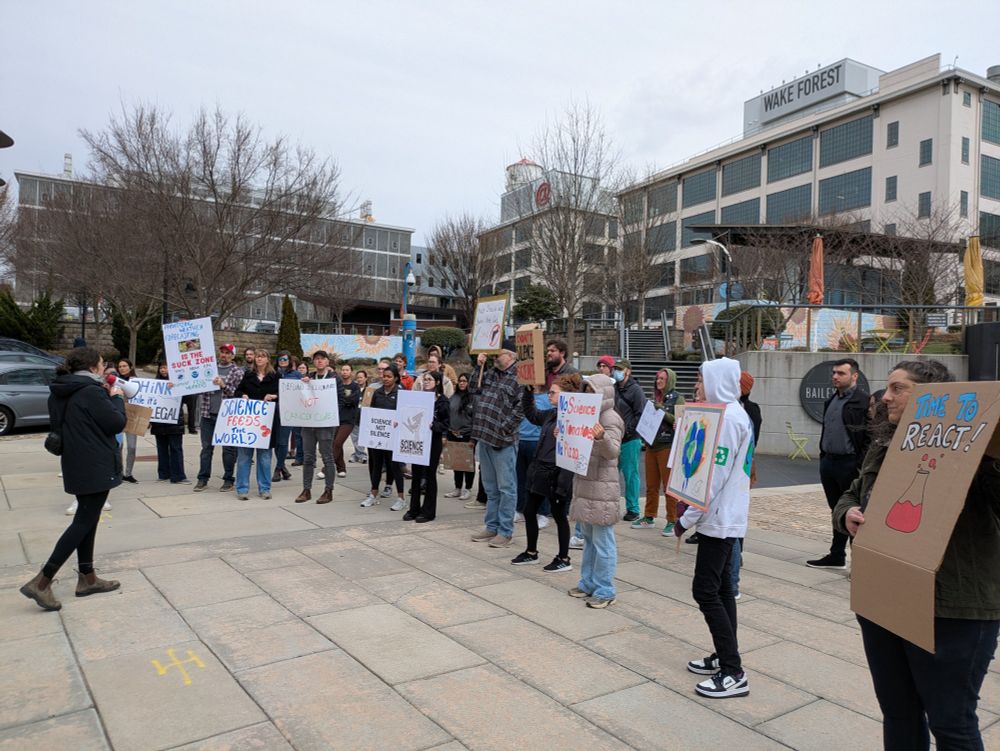 Dozens of people protesting the current political attacks on scientific research. They are gathered together in a local park, holding protest signs and listening to group members speak via megaphone.