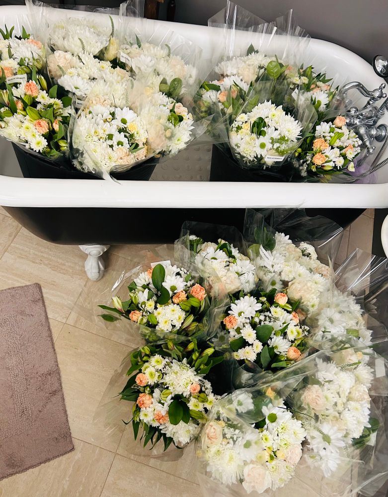 Flowers sitting in black gardening tubs set in a clawfoot bathtub and on the tile floor of a bathroom.