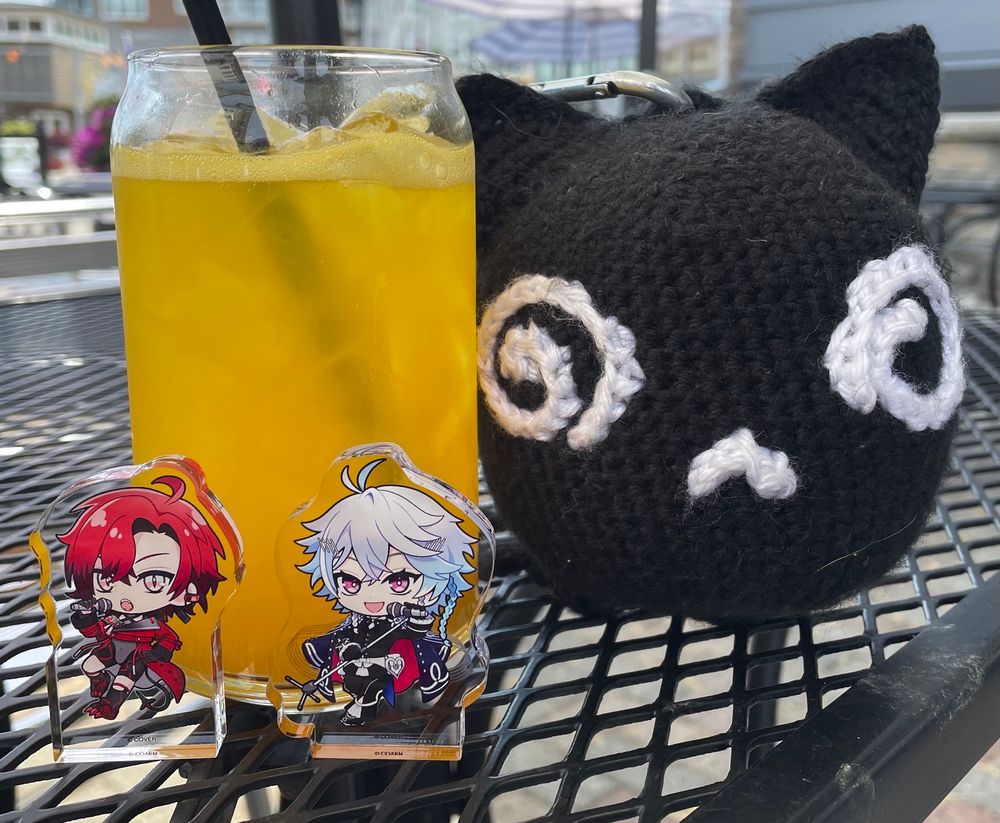 Acrylic block standees of Holostars EN VTubers Machina X Flayon and Octavio standing in front of a mango drink. To the right is a crocheted Machiroon plush
