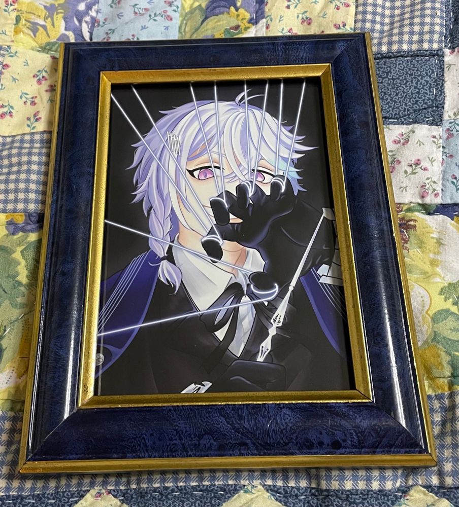 A photo of a picture frame laying on a quilt. The frame is dark blue with gold accents. In the frame is a print of Holostars EN Vtuber Octavio, drawn by Renrolo. Octavio is holding a baton in one hand and pulling puppet strings in the other.