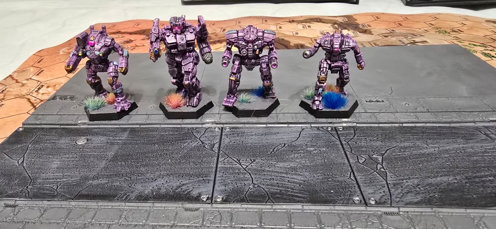 Four battletech miniatures painted in purple and teal, displayed on a roadside terrain plate.  The mechs pictured are a Charger, an Assassin, an ostroc, and a Merlin.