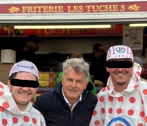 Fabien Roussel durant le tour de France
en avant : Roussel en kway noir, entre 2 personne portant des ponchos et des casquettes Leclerc
en arriere plant : une friterie LES TUCHES 3