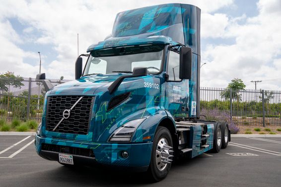 A bright blue zero-emission Volvo semi-truck is parked in an outdoor lot. The truck features futuristic graphics and the words “Zero Emission” and “Los Angeles” on the side. It's a cab-only model with no trailer attached, and a tall fence and trees line the background under a partly cloudy sky. 