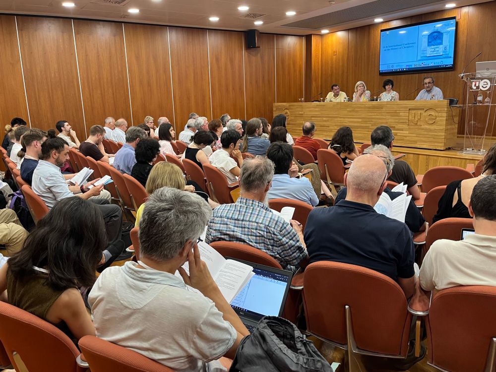 A photograph of an auditorium from the back of the room in which an audience is seen watching a four-person panel on the stage