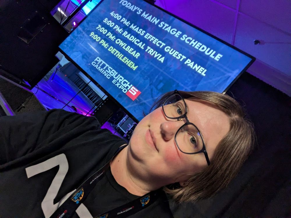 A white person with short brown hair and glasses standing in front of a television displaying a main stage schedule, with the Mass Effect guest panel at 4pm.