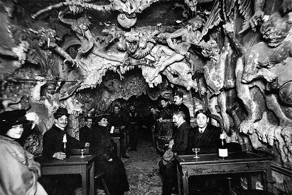 A black-and-white photo of patrons, mostly smiling, sitting at table in the interior of a bar designed to look like a cave with sculpted demons and monsters covering the ceiling