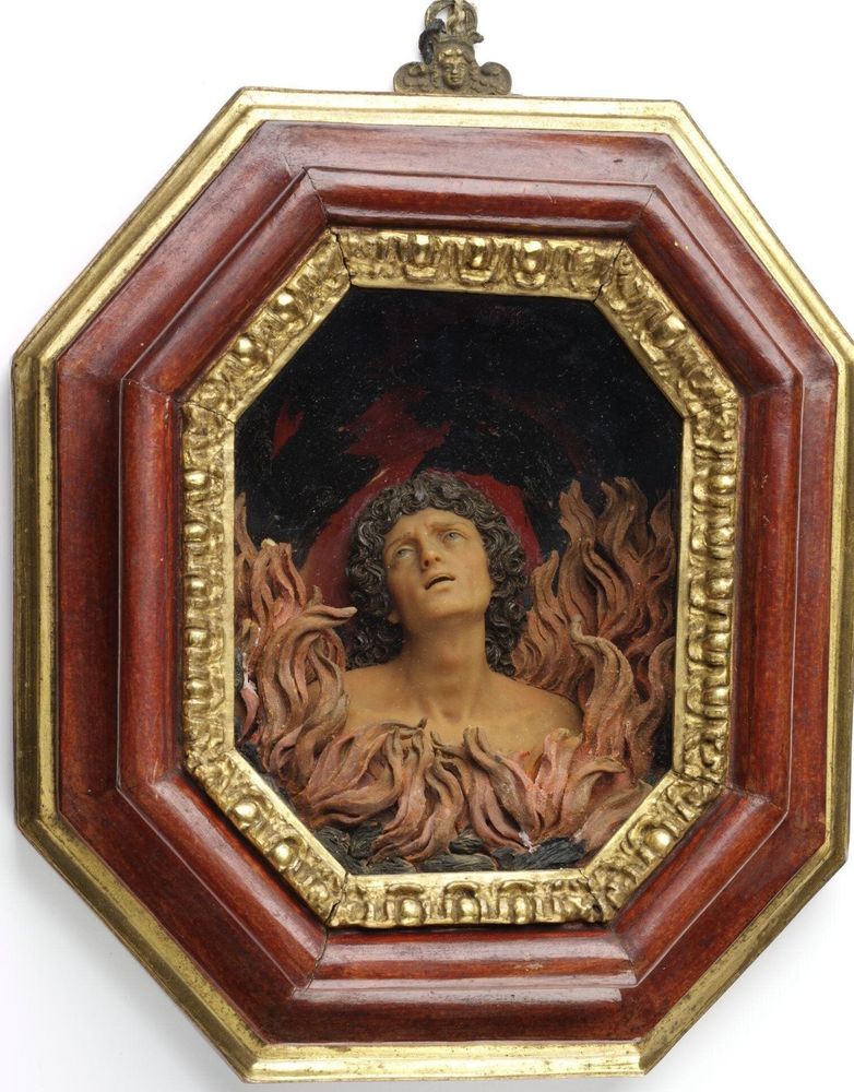 "A naked male bust emerging from modelled flames and coals. The background and sides of the interior of the deep stained and gilded box frame are painted red and black to resemble flames. The head of the ‘soul’ has long curly hair and his eyes are cast upward in supplication with his mouth is open."