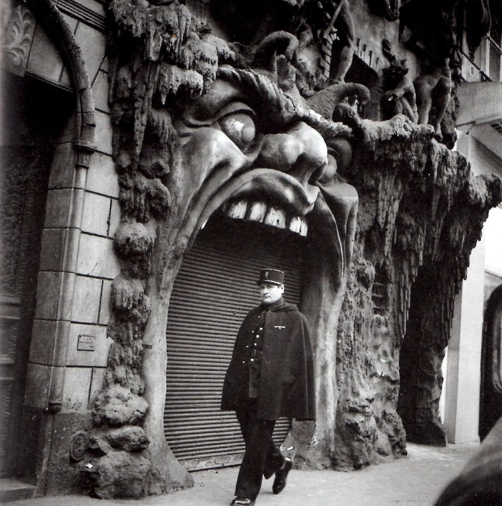 A black-and-white photo of a gendarme walking past a doorway shaped like a gaping Hell mouth