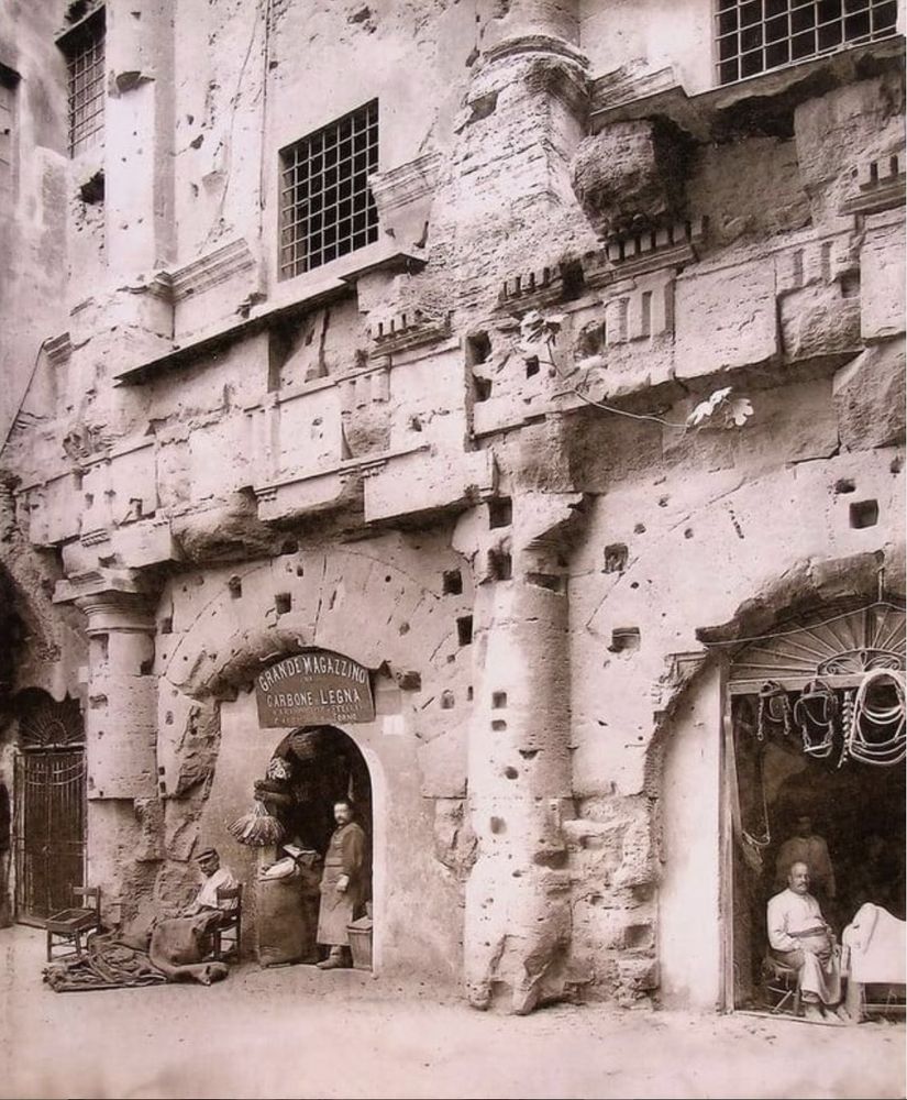 A sepia photograph showing part of the Theater of Marcellus in Rome. Two men are in an archway with a sign reading "Grande Magazzino di Carbone di Legno," the other with a man seated with bridles (?) hanging over the entryway