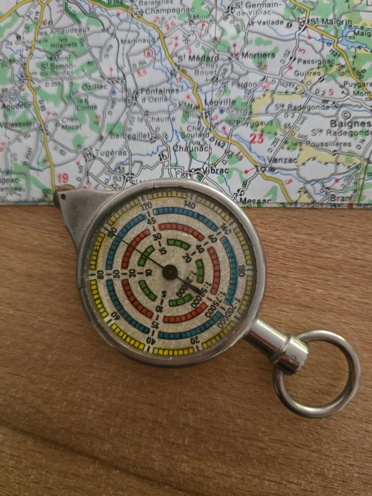 Auf einer Holzfläche liegt eine Landkarte. Davor liegt ein alter Kilometerzähler aus Metall. Auf einer runden Scheibe sind vier verschiedene Maßstäbe als Skalen eingezeichnet. Darüber liegt ein schwarzer Zeiger. Oben ist ein kleiner Metallstab mit eingelassenem Ring als Griff, unten läuft er spitz zu und endet in einem Rädchen. 