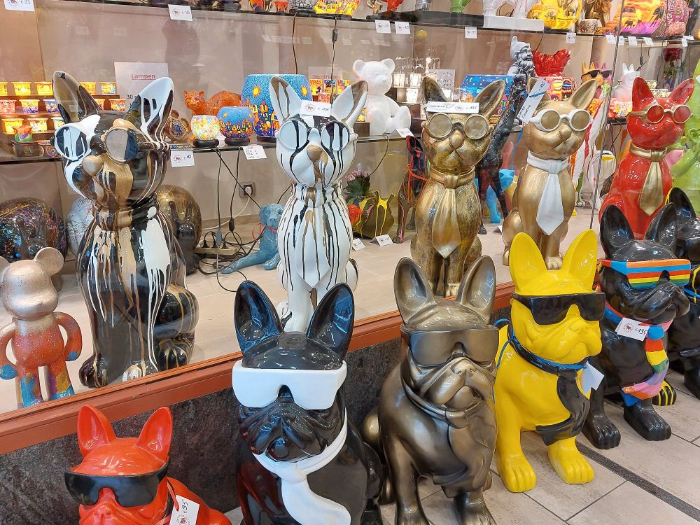 In einem Schaufenster stehen diverse knallbunte Figuren. Ganz vorn sitzt eine Reihe großer Katzen mit Brille. Sie sind goldfarben oder farbig mit Gold. Vor dem Schaufenster sitzt eine entsprechende Reihe mit Hunden. Sie tragen zur Brille noch einen Schal.