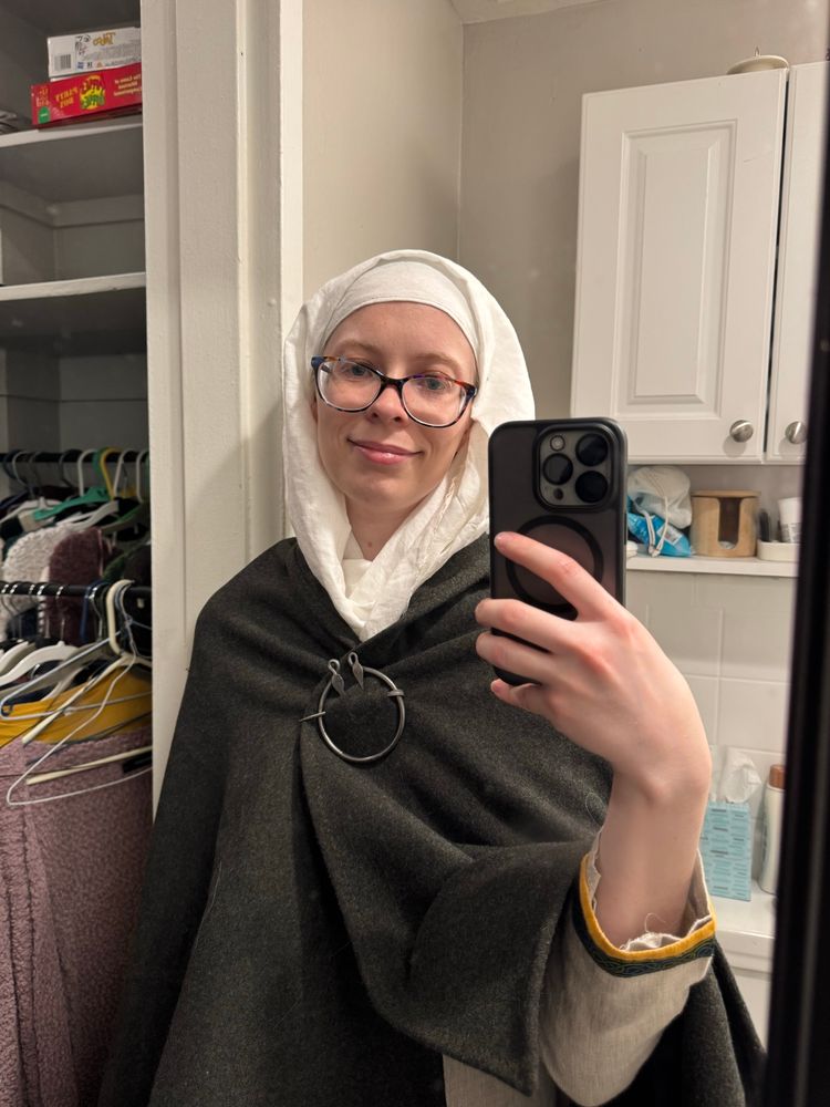 Awkward bathroom selfie of me in 10th century dress, a white veil and a dark green cloak over a gray dress. Yes the glasses are anachronistic but I can’t see without them.