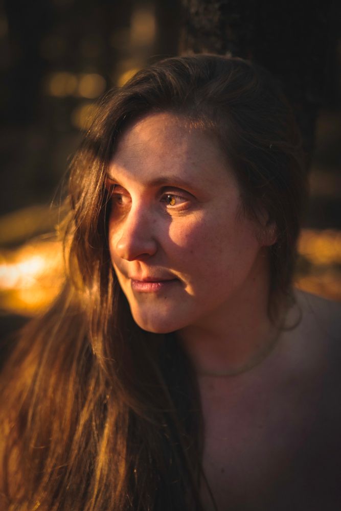 close up photo of the face and shoulders of Jessy, a white gender fluid person with Hazel eyes and waist length brown hair. They have a calm content smile as the amber sun warms their face, distantly contemplating a world far from this cozy forest groove on a perfect autumn day.