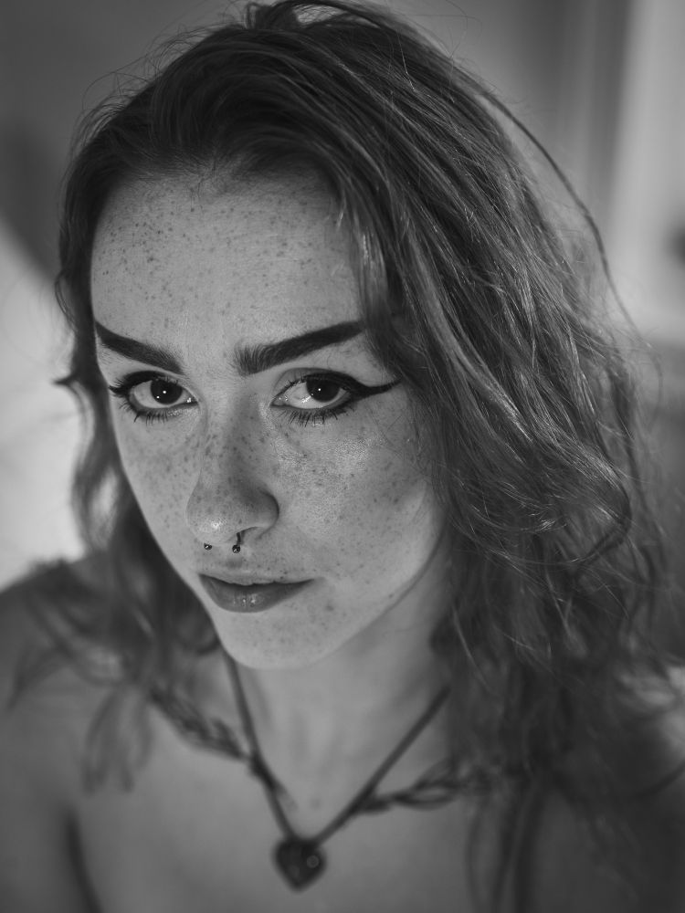 Myah is a white queer woman with long hair and freckles, wing tip eye makeup, a septum ring, and is wearing a little heart necklace. Image is taken from above with them looking up at the camera