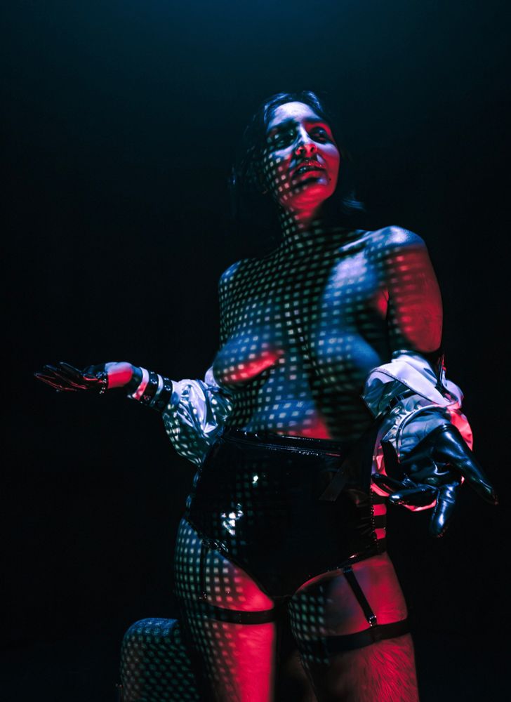 Image of a non binary Punjabi model with medium dark skin, medium length black hair, and glittery lipstick, wearing an open long sleeve opalescent bolero jacket, and black strapped booty shorts. They are in a black void lit in blue and red saturated light textured by white square Pixels projected on their skin. They lower their open jacket down to their elbows exposing their shoulders and chest in a gesture of nonchalance.