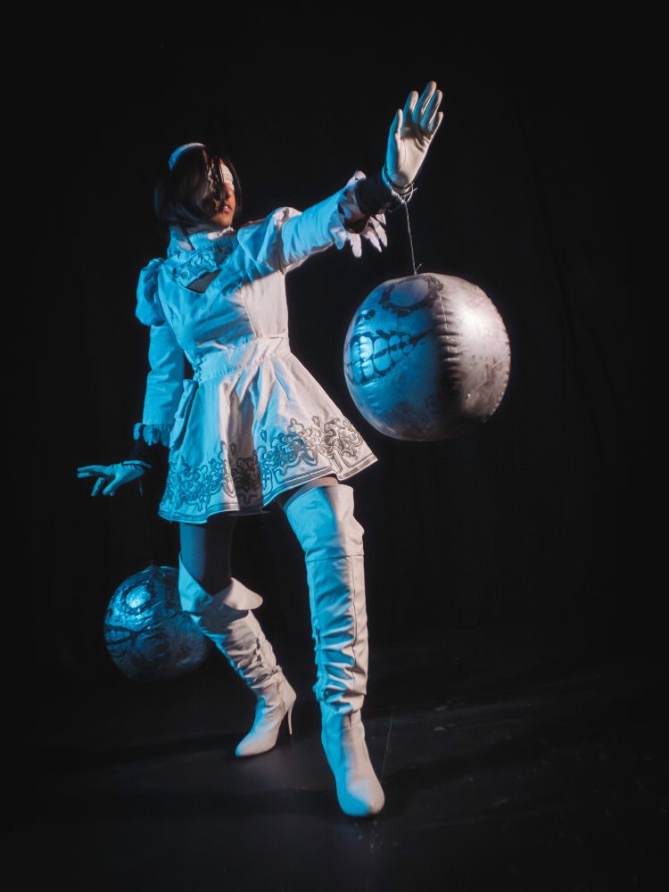 a non binary cosplayer with brown hair and medium dark skin, dressed as the character 2P from the Neir:Automata spin-off media.  Costume consists of a white dress with embroidered grey patterns, tall white boots, and white gloves and blindfold. Floating next to them are two spherical skulls modelled after the character Emil from the same game. They are lit by white light from one side and cool blue from the other, standing against a black background