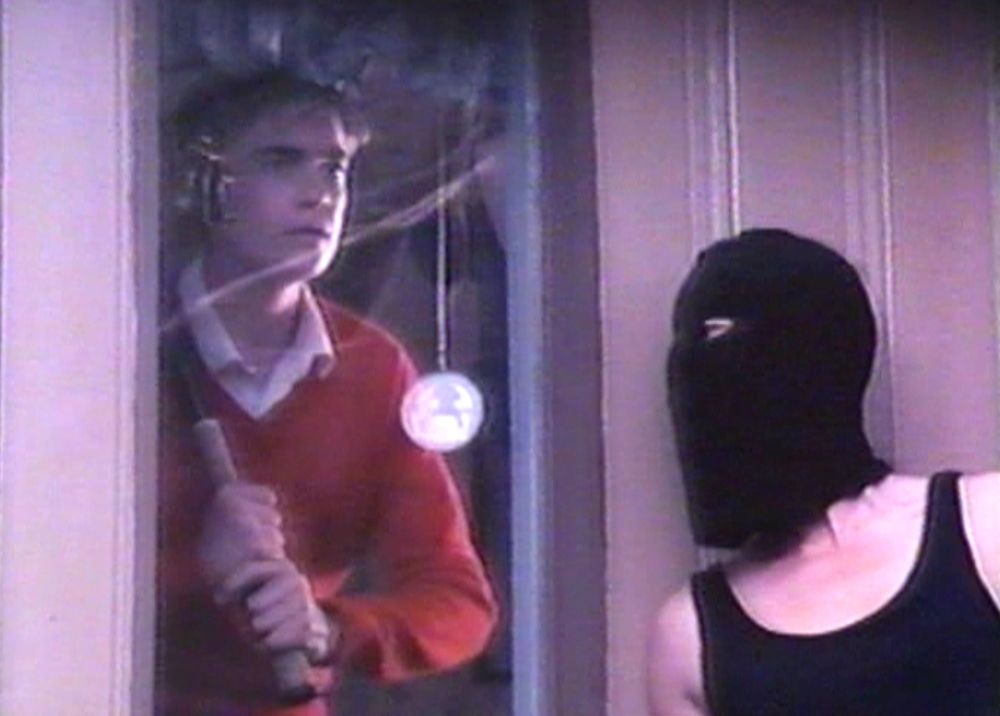 Troy McGreggor with baseball bat in hands, looks out of a window of his aunt's house, while an evil cult member hides against a wall in an image from the hit 1990 film The Final Sacrifice.