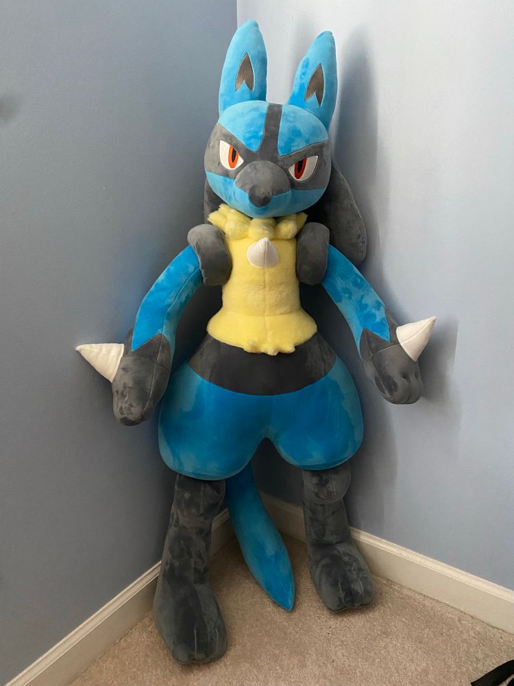 A photograph of a life-sized Lucario plush, standing in the corner of a room against a blue-ish grey wall. 