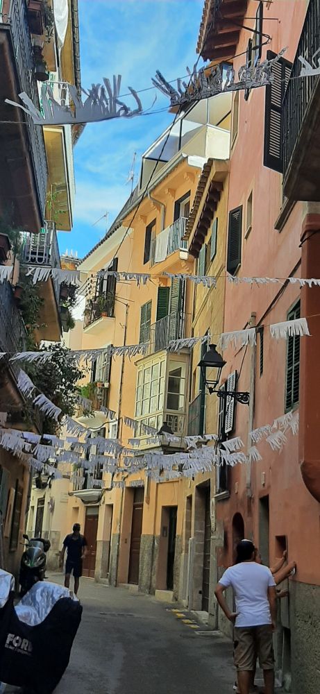Eine kleine Gasse (Carrer Contramoll Mollet) irgendwo auf Palma: Beige Hauswände, trotz Sonne schattig, sich unterhaltende Spanier, Flatterbänder zwischen den Fassaden, die nur drei bis maximal vier Meter auseinanderliegen — gefühlt zu schmal für ein Pkw...