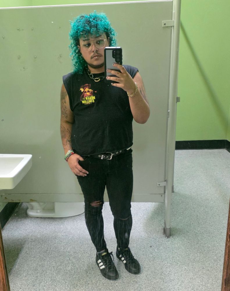 levi taking a picture in a bar bathroom mirror. hes a Black trans man with curly deep teal hair cut into a mullet, wearing all black with a Megan thee Stallion shirt. 