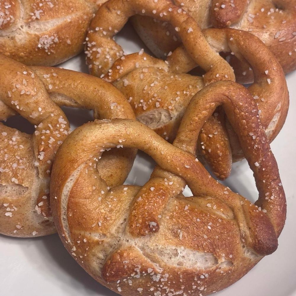 Closeup photo of a plate of soft Bavarian-style pretzels. They are golden in color with darker arms and broad, fluffy middle. There are about 5 seen on the white plate