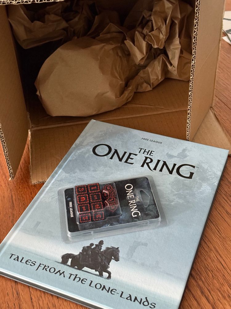 A photo of The One Ring second edition book, “Tales From The Lone-Lands” and a set of black and red the one ring dice. 