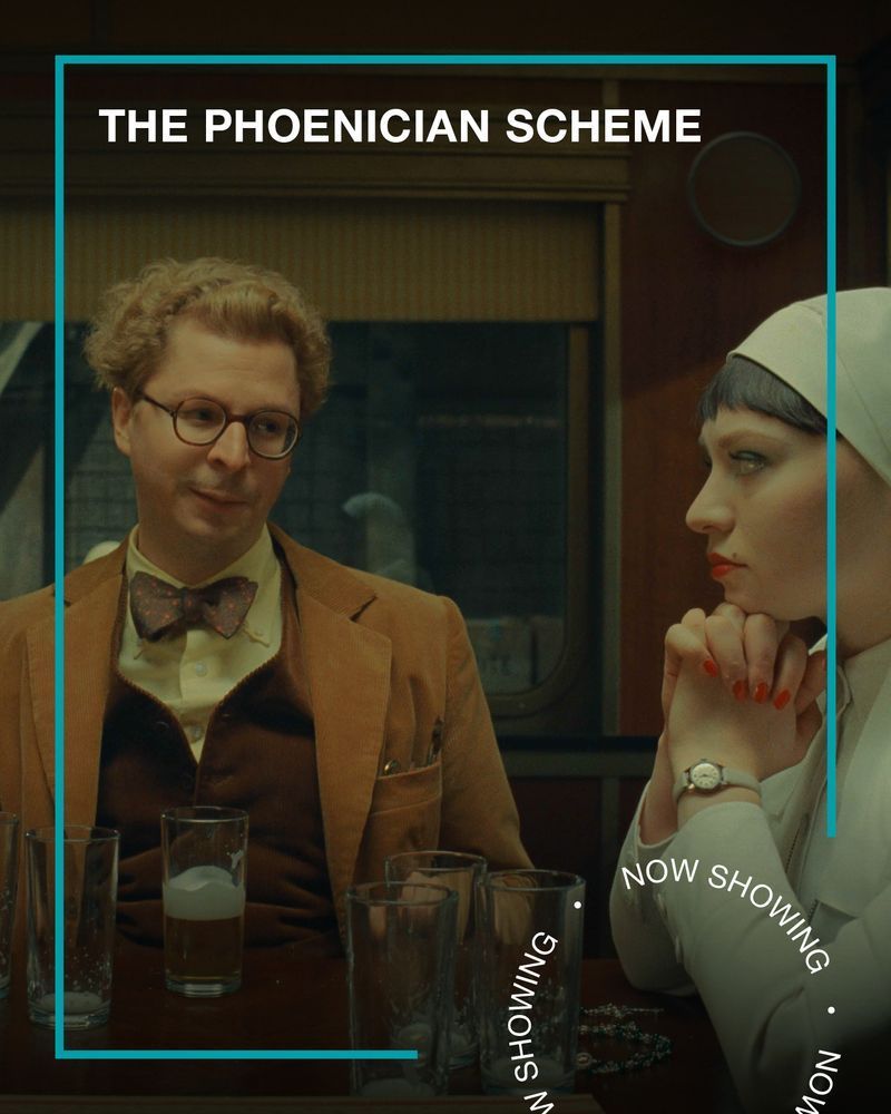 A man in a camel suit, glasses and a bow tie smiles at a nun dressed in white. Text - The Phoenician Scheme, now Showing.