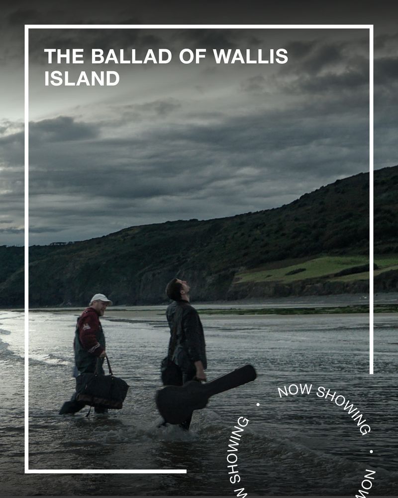 Two men, fully dressed, stand in shallow sea water holding guitar cases. Text - The Ballad of Wallis Island, Now Showing.
