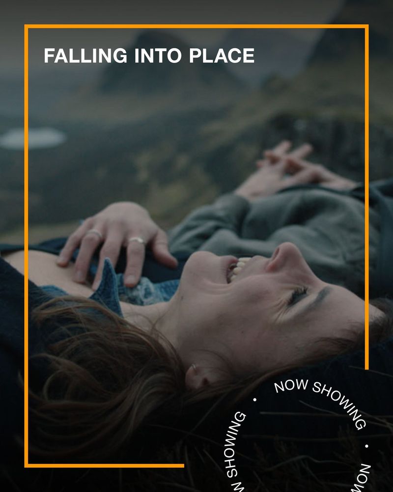 Two people lie on a hill, laughing. Text - Falling Into Place, Now Showing.