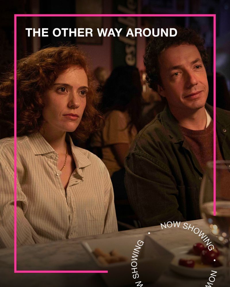 A man and a woman sit next to each other at a dinner table. Text - The Other Way Around, Now Showing.
