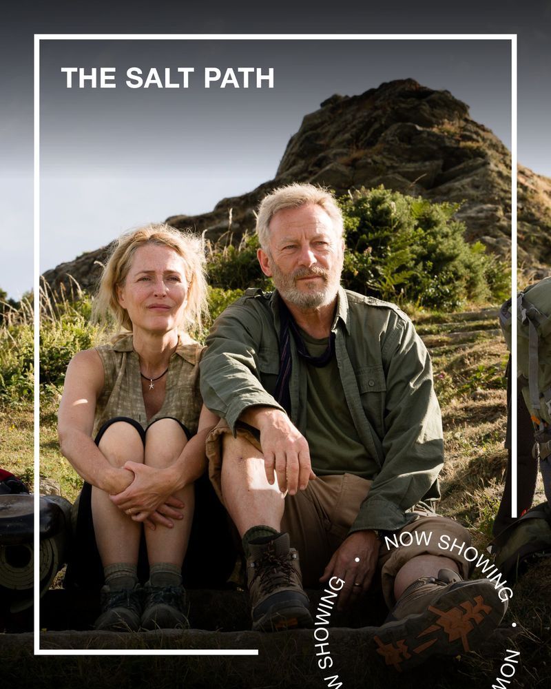Two people wearing hiking clothing sit on a sunny hill. Text - The Salt Path, Now Showing.