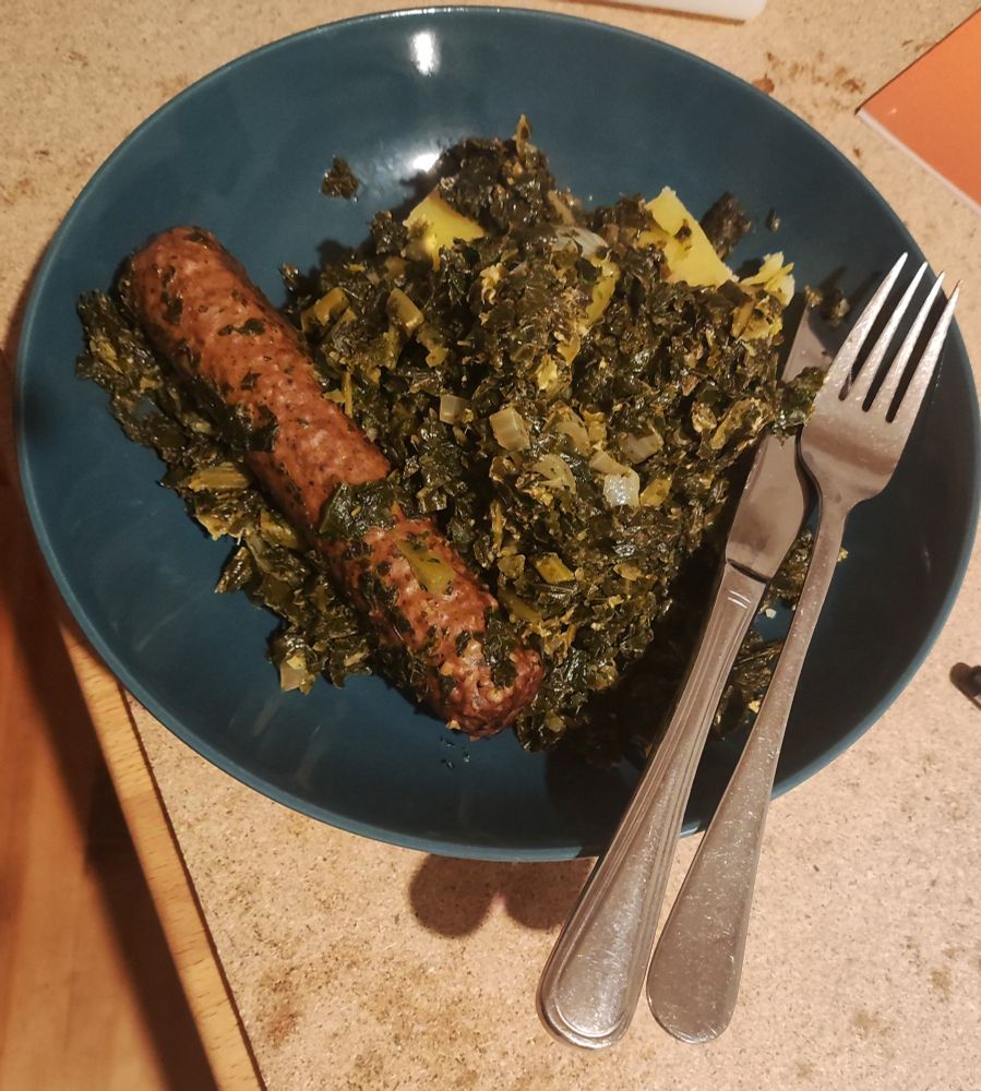Eine Portion Grünkohl auf Kartoffeln und dazu ein leckerer veganer Pinkel. Das ganze auf einem Petrolfarbenem Teller mit Messer und Gabel.