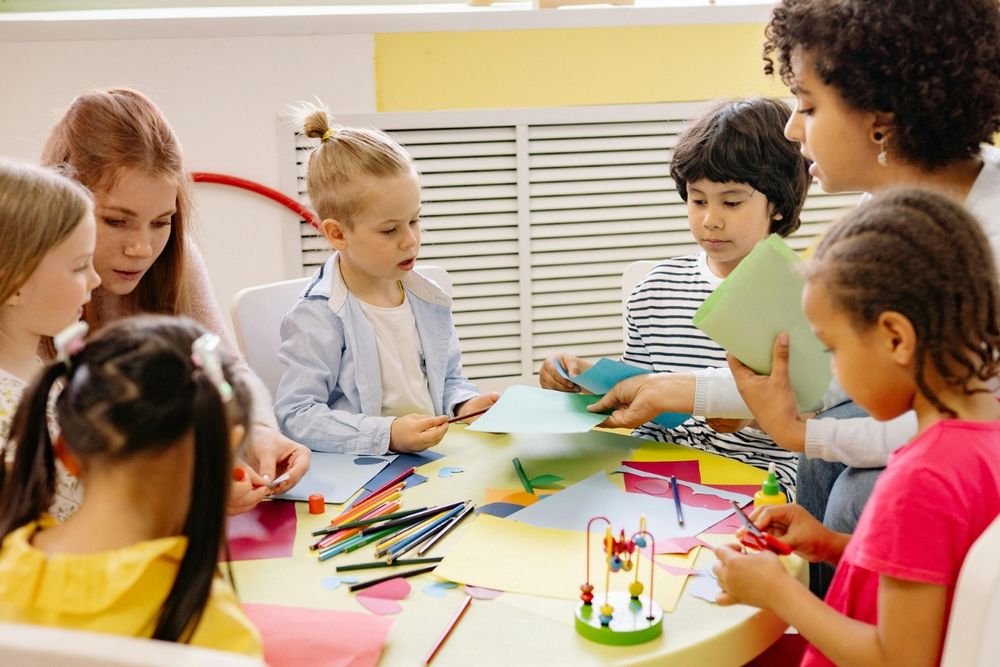 A group of young children sit around a table with two adults, engaged in an arts and crafts activity using coloured paper, pencils, and glue in a bright classroom setting.