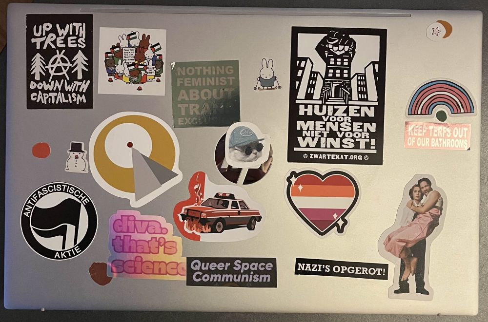 a silver laptop shown closed above. The stickers are: a black and white print with the anarchist symbol inbetween two trees, reading: up with trees, down with capitalism. A collection of miffy characters wearing kuffiye and holding palestinian flags. A holo lettering that reads: nothing feminist about trans exclusion. Miffy on skis. A drawing of a block of houses, the middle shaped like a fist, with the caption "homes for people, not profit" (translated from dutch). A trans rainbow. A holo lettering that reads: keep terfs out of our bathrooms. A snowman wearing a top hat. The Vulcan "infinite diversity in infinite combinations" symbol from Star Trek. The face of a cat wearing a cap and round sunglasses. A sparkling heart emoji with lesbian flag colours. A circle with two black flags and the caption "antifascist action" (translated from german). A holo lettering that reads: "Diva. that's science." A drawing in red and white tones of a cop car on fire. A lettering that reads Queer Space Communism. A lettering that reads nazi's fuck off (translated from dutch). And lastly, Gillian Anderson wearing a suit, lifting David Duchovny wearing a pink sleeveless dress like he's a kid. 