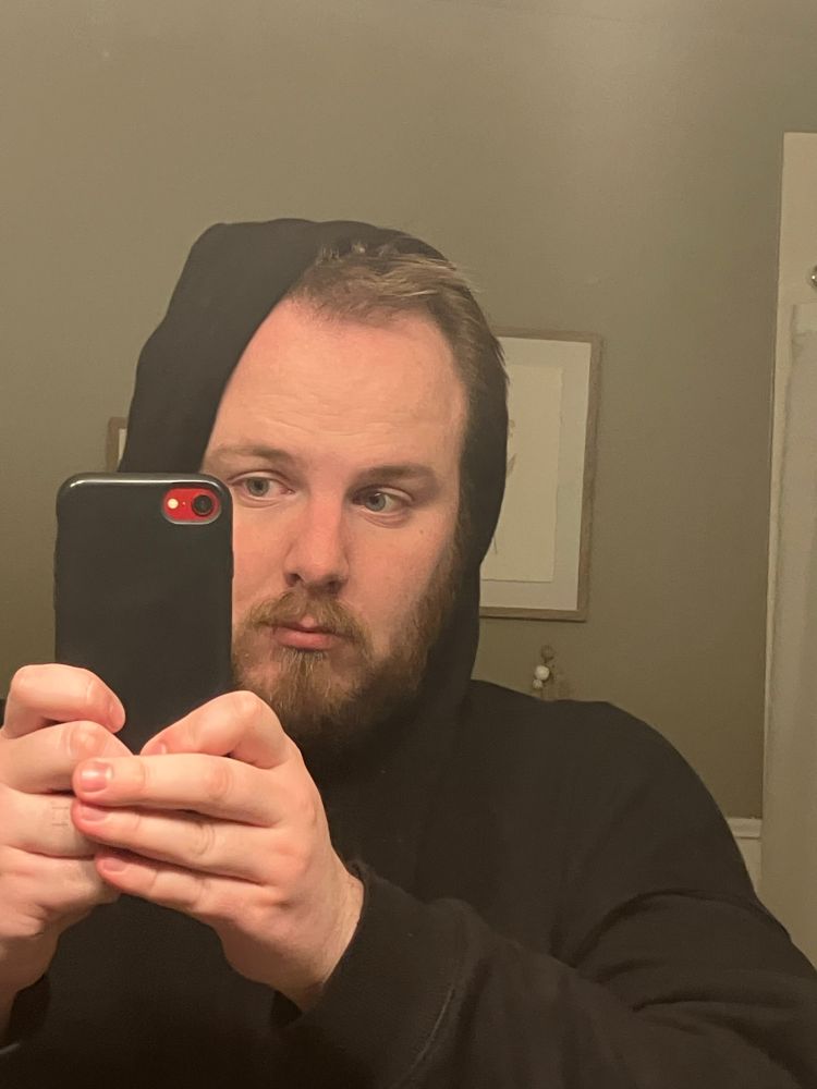 Nick, who is white with a beard, stand in front of mirror in a black hoodie with his phone in his hand.