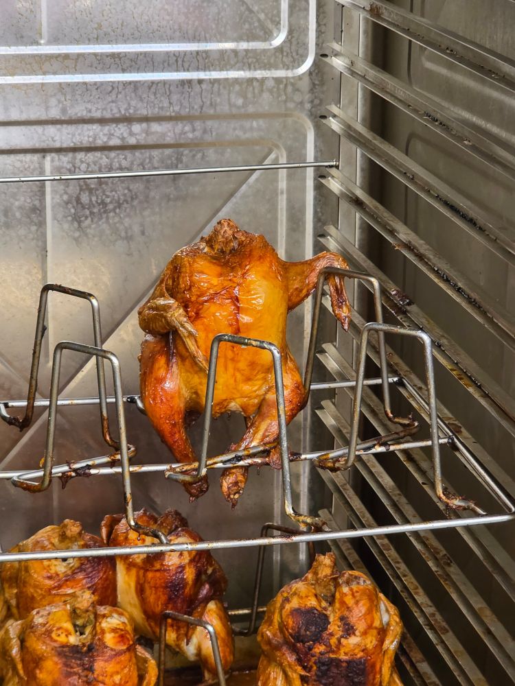 Photo of a rotisserie chicken sitting upright on a rack in a commercial oven. The chicken's wing is hooked around the adjacent rack and it gives the impression that the chicken is lounging all chill