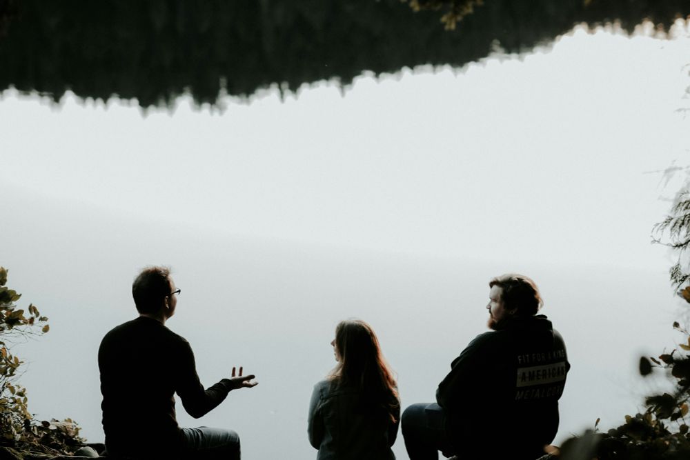 A black-and-white photograph; the upper portion of which is dominated by the clear space of a large body of water, with a treeline reflected at the very top. Along the bottom of the photo, a man sits with another man and a woman, gesturing with his hand as he talks to them.