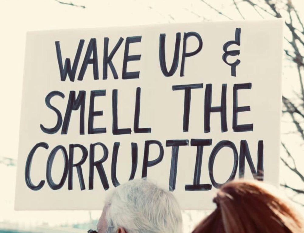A handmade protest sign reading "WAKE UP & SMELL THE CORRUPTION."