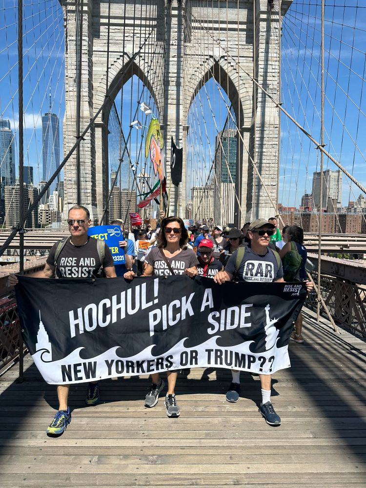 Three members of Rise and Resist marching across the Brooklyn Bridge with a banner that reads “Hochul! Pick a side: New Yorkers or Trump?