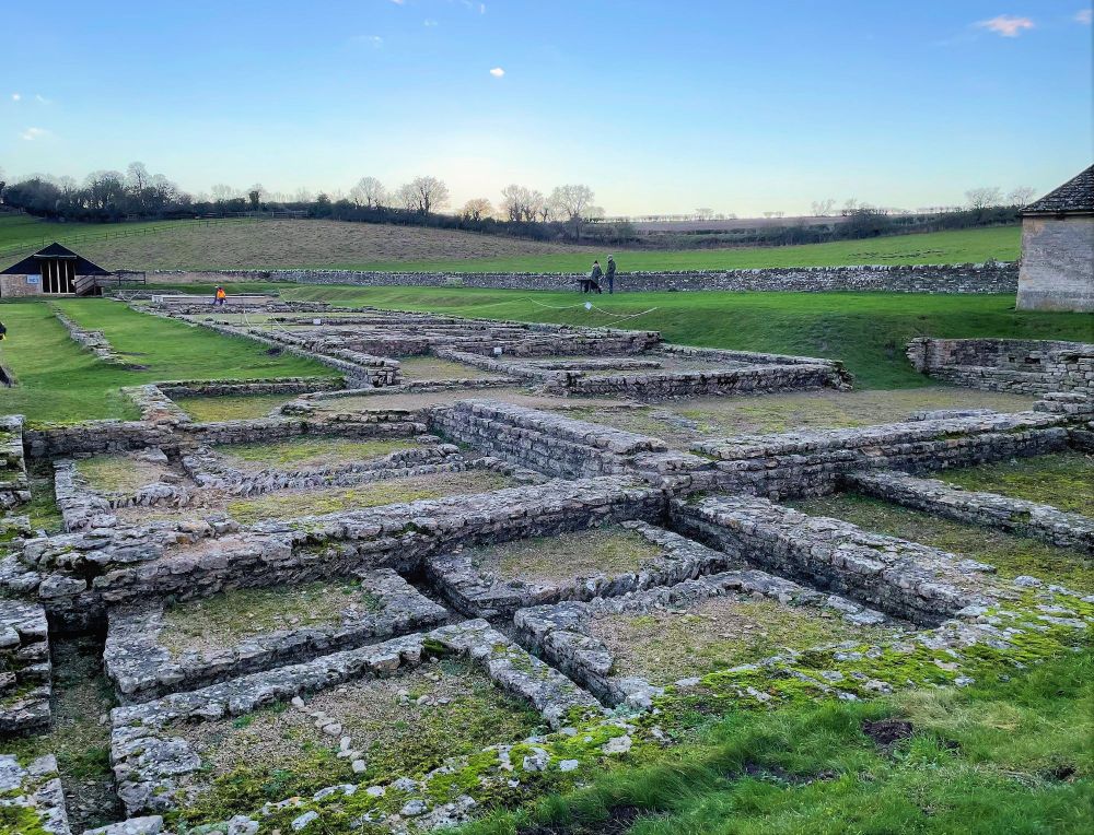 View across the site, taken on a cold clear winter's day. A cross-shaped channel hypocaust is in the foreground, while low stone walls forming the footings of the west wing stretch away to a modern mosaic cover building.