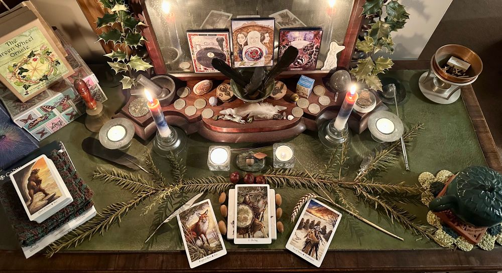 A wooden desk with a green leather top is covered in candles, feathers, greenery, tarot cards, oracle cards and various other totems and crystals. 