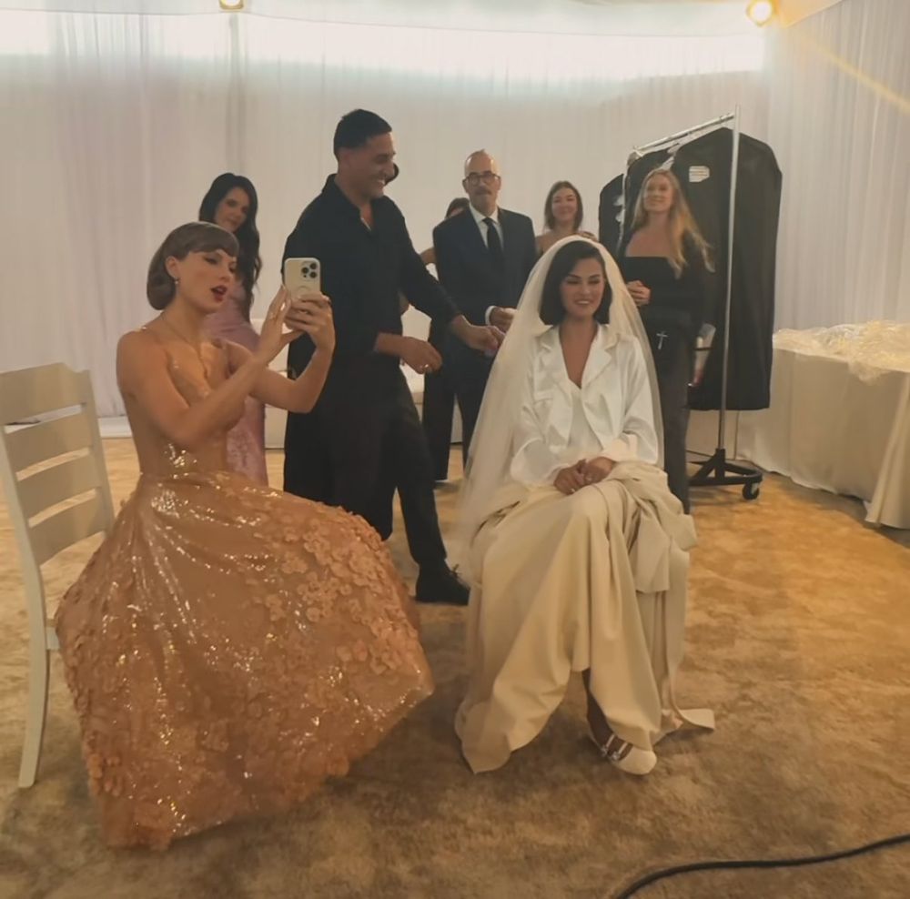 Taylor Swift and Selena Gomez, at what looks like Selena’s dressing room for her wedding. They are both sitting down. Taylor is holding up her phone talking a mirror selfie. She is wearing a floor lenght gown in a beige/ peach color, with flower detailing towards the end of the dress.  Selena is wearing what looks a like white silk pj set and a veil. 