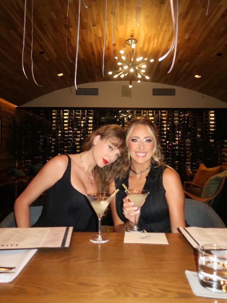 Taylor Swift and Brittany Mahomes. They are in what looks lime a restaurant. Both of them are wearing black shirts. Taylor is wearing a black corset, and a “T” necklace with a gold chain and red stones. 