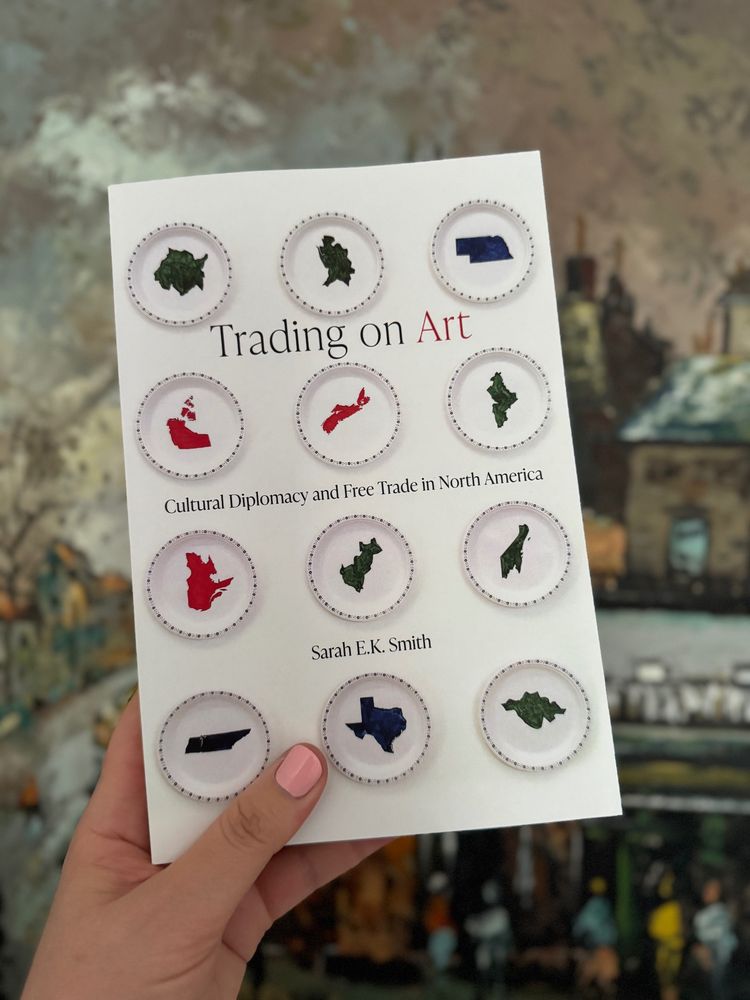 A hand with pink nail polish holds a book titled Trading on Art: Cultural Diplomacy and Free Trade in North America by Sarah E.K. Smith. The book cover shows twelve paper plates, on each a state or province from Canada, Mexico and the US is rendered in red, blue or green. In the background you can see the brushstrokes of a grey painting with a building and people.