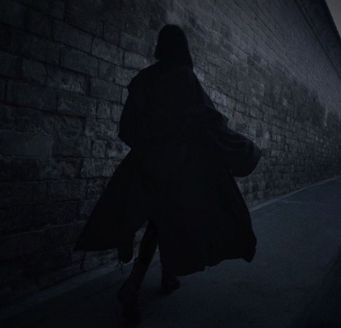 A photograph of a figure in a black cloak running down an empty alley way at night  